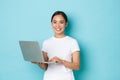 Portrait of smiling beautiful asian girl standing over light blue background in white casual t-shirt, using laptop Royalty Free Stock Photo