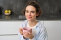 Portrait of smiling, beautiful young woman, drinking coffee in kitchen, morning magic with cuppa tea, looking tenderly Royalty Free Stock Photo