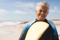 Portrait of smiling biracial senior woman carrying surfboard at beach against sky on sunny day Royalty Free Stock Photo