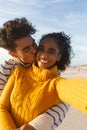 Portrait of smiling biracial woman taking selfie of boyfriend hugging and kissing on cheek at beach Royalty Free Stock Photo