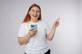 Portrait of a smiling casual woman holding smartphone and pointing finger away at copy space over white background. 30s Royalty Free Stock Photo