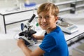 Portrait of smiling caucasian elementary schoolboy with microscope sitting at desk in laboratory Royalty Free Stock Photo