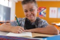 Portrait of smiling caucasian schoolboy with face mask sitting in classroom, writing in schoolbook