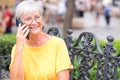 Portrait of smiling caucasian senior woman in yellow t-shirt and glasses sitting outdoors on a bench talking on mobile phone Royalty Free Stock Photo