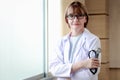 Portrait of smiling confident female doctor in white workwear wearing glasses, holding stethoscope, standing with arms crossed by Royalty Free Stock Photo