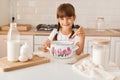 Portrait of smiling cute little girl baker with braids on kitchen with baking ingredients, looking at camera with charming smile, Royalty Free Stock Photo