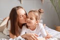 Portrait of smiling cute little girl lying in bed with mother, woman kissing her daughter with love Royalty Free Stock Photo