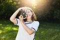 Portrait of smiling cute woman wearing white blouse taking photo with her retro camera while standing in green park or on meadow. Royalty Free Stock Photo