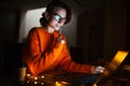 Portrait of smiling designer girl, working at graphic tablet on laptop. Wearing eyeglasses and orange sweater. In dark room home. Royalty Free Stock Photo