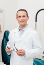 portrait of smiling doctor in white coat with eyeglasses in hands looking at camera