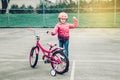 smiling excited Caucasian preschooler girl standing with pink bicycle in helmet on court field outside on spring day showing champ Royalty Free Stock Photo