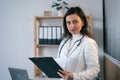 Portrait of smiling female doctor with lab coat in her office holding a clipboard with medical records looking at camera Royalty Free Stock Photo