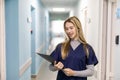 Portrait Of Smiling Female Doctor Wearing Scrubs Standing In Hospital Corridor Holding Clipboard Royalty Free Stock Photo