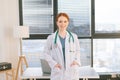 Portrait of smiling female doctor in white coat standing on background of window in sunny day in light medical clinic Royalty Free Stock Photo