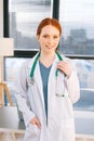 Portrait of smiling female doctor in white coat standing on background of window in sunny day in light medical clinic Royalty Free Stock Photo