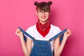 Portrait of smiling female with funny bunches, wears t shirt, denim overalls and bandana on neck, has toothy smile, pulls