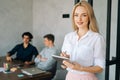 Portrait of smiling female office worker in casual clothes taking notes in her diary standing in office, looking at Royalty Free Stock Photo