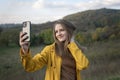 Portrait of smiling girl taking selfie on smartphone on mountain landscape background. Young woman in yellow park jacket enjoys