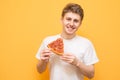 Portrait of a smiling guy dressed in a white T-shirt isolated on a yellow background, holding a piece of pizza in his hands and Royalty Free Stock Photo