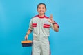 Portrait of smiling handsome man wearing mechanic uniform holding tool box and showing gesturing okay sign isolated on blue