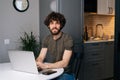 Portrait of smiling handsome young man using laptop sitting at table in kitchen room with modern interior, looking at Royalty Free Stock Photo