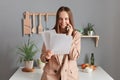 Portrait of smiling happy brown haired businesswoman wearing beige suit standing near table on kitchen at home, holding documents Royalty Free Stock Photo