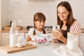Portrait of smiling happy mother baking together with her daughter, sitting at table in the kitchen and using smart phone, finding
