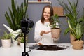 Portrait of smiling happy satisfied Caucasian woman botanist showing flowerpot with green plant to camera on tripod, recording