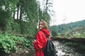 Portrait of smiling hiker girl hiking, looking into camera and smiling on stream and mountains background, wearing red raincoat.