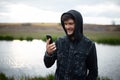Portrait of smiling hooded man, looking in smartphone, outside on background of river.