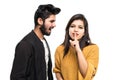 Portrait of a smiling indian couple, man quarrel but woman showing silence gesture and looking at camera over gray wall