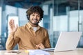 Portrait of a smiling Indian young man wearing a suit sitting in the office at a desk with a laptop, looking and waving Royalty Free Stock Photo