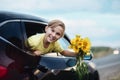 Portrait of smiling kid boy teen look from the car holding bouquet of flowers. Holiday, travel and adventure concept