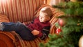 Portrait of smiling little boy lying on sofa next to beautiful Christmas tree in living room and palying on digital