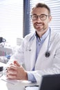 Portrait Of Smiling Male Doctor Wearing White Coat With Stethoscope Sitting Behind Desk In Office Royalty Free Stock Photo