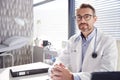 Portrait Of Smiling Male Doctor Wearing White Coat With Stethoscope Sitting Behind Desk In Office Royalty Free Stock Photo