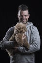 Portrait of a smiling man looking at the camera with a small red poodle dog. Black background. Vertical Royalty Free Stock Photo