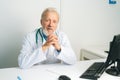 Portrait of smiling mature adult male doctor in white coat with stethoscope sitting at desk with computer in medical Royalty Free Stock Photo