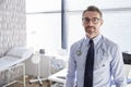 Portrait Of Smiling Mature Male Doctor With Stethoscope Standing By Desk In Office Royalty Free Stock Photo