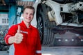 Portrait of smiling mechanic in red uniform giving thump up near lifted vehicle, auto mechanic check wheel at garage, technician