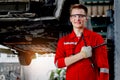Portrait of smiling mechanic in red uniform standing and holding tool near lifted vehicle, auto mechanic check wheel at garage,
