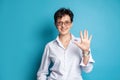 Portrait of smiling middle adged woman in glasses showing five fingers against blue studio wall background Royalty Free Stock Photo