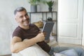 Portrait of smiling middle aged man using digital tablet pc sitting on the sofa in home interior. Grey haired freelancer Royalty Free Stock Photo