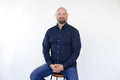 Portrait of smiling middle-aged man wear blue shirt, jeans sitting on stool, interlacing fingers on white background.