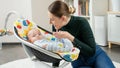 Portrait of smiling mother sitting next to little baby son rocking in electric chair. Child development and happy