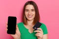 Portrait of smiling optimistic dark haired woman holding credit card and cell phone with black black screen for advertisement,