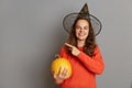 Portrait of smiling optimistic young adult happy woman in black witch hat holding pumpkin in hands, pointing aside at copy space Royalty Free Stock Photo