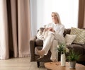 Portrait of smiling pleasant old mature blond woman sitting on sofa with handmade clothes in hands