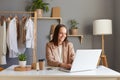 Portrait of smiling positive brown-haired woman wearing beige jacket sitting at her workplace in front of laptop, typing on Royalty Free Stock Photo
