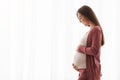 Portrait Of Smiling Pregnant Woman Tenderly Embracing Belly Against Window At Home Royalty Free Stock Photo